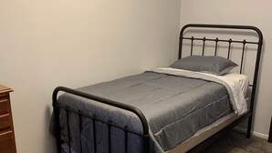 Un lit simple avec un cadre en métal et une literie grise, dans une pièce dont le sol est recouvert d’une moquette et qui contient une commode en bois.