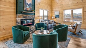 Interior acogedor de una cabaña de troncos con chimenea de piedra, sillones tapizados en verde, un sofá y un ventanal con persianas.