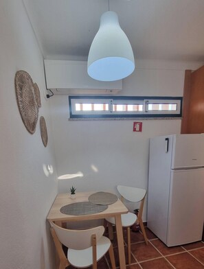 Eine kleine Küche mit einem weißen Kühlschrank, einem Tisch mit Stühlen, einer Hängelampe und einem Fenster mit Jalousien.
