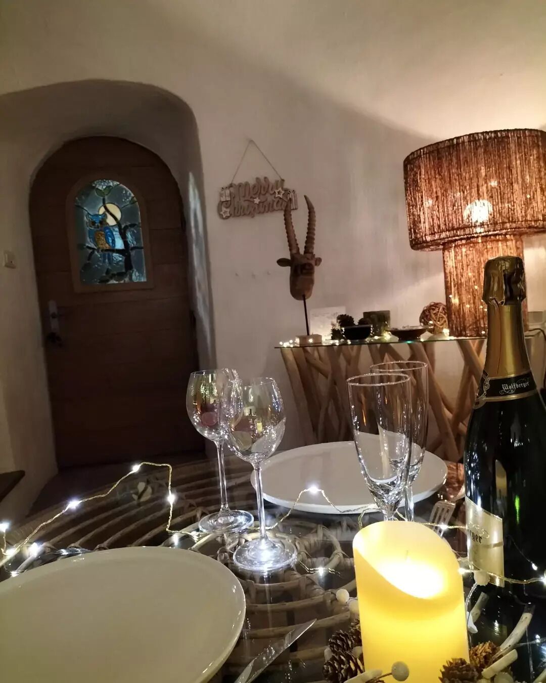 Una mesa de comedor preparada con copas de vino, platos, una vela y una botella de vino, con un detalle decorativo en la pared y una puerta al fondo.