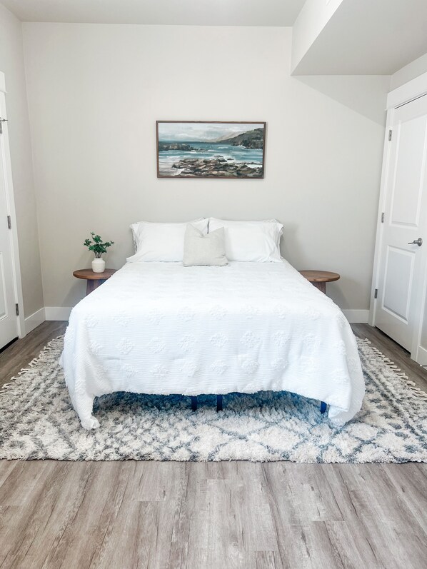 Un dormitorio con una cama, un cuadro, una mesita con una planta y una alfombra.