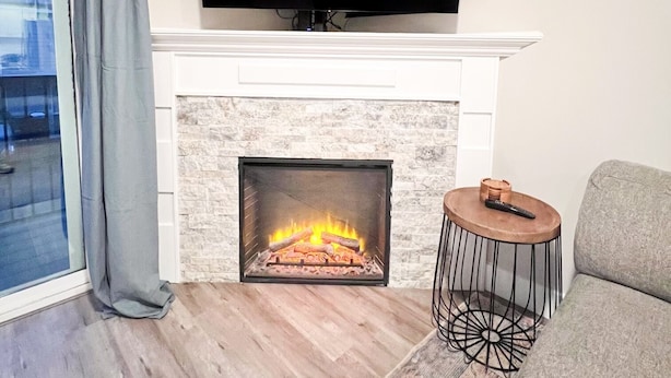 A living room with a fireplace, a flat-screen TV mounted above it, a grey sofa, and a small round side table with a remote control. - Beautiful, newly built 4 bedroom with garage (Yakima)