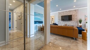 A modern living room with a large sectional sofa, a flat-screen TV mounted on a brick wall, and a glass partition leading to another room. - Renovated Beachfront 3 BR - Steps to Baha Mar (Nassau)