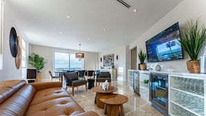 A modern living room with a leather sofa, wooden coffee table, and dining area with chairs. A large TV is mounted on the wall, and there are potted plants. - Beachfront 3BR Penthouse w/Terrace Next to BahaMar (Nassau)