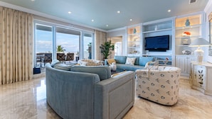 A spacious living room with a large sofa, a coffee table, and a TV. There are shelves with decorative items and a dining area with a table and chairs. - Beachfront 3BR Penthouse w/Terrace Next to BahaMar (Nassau)