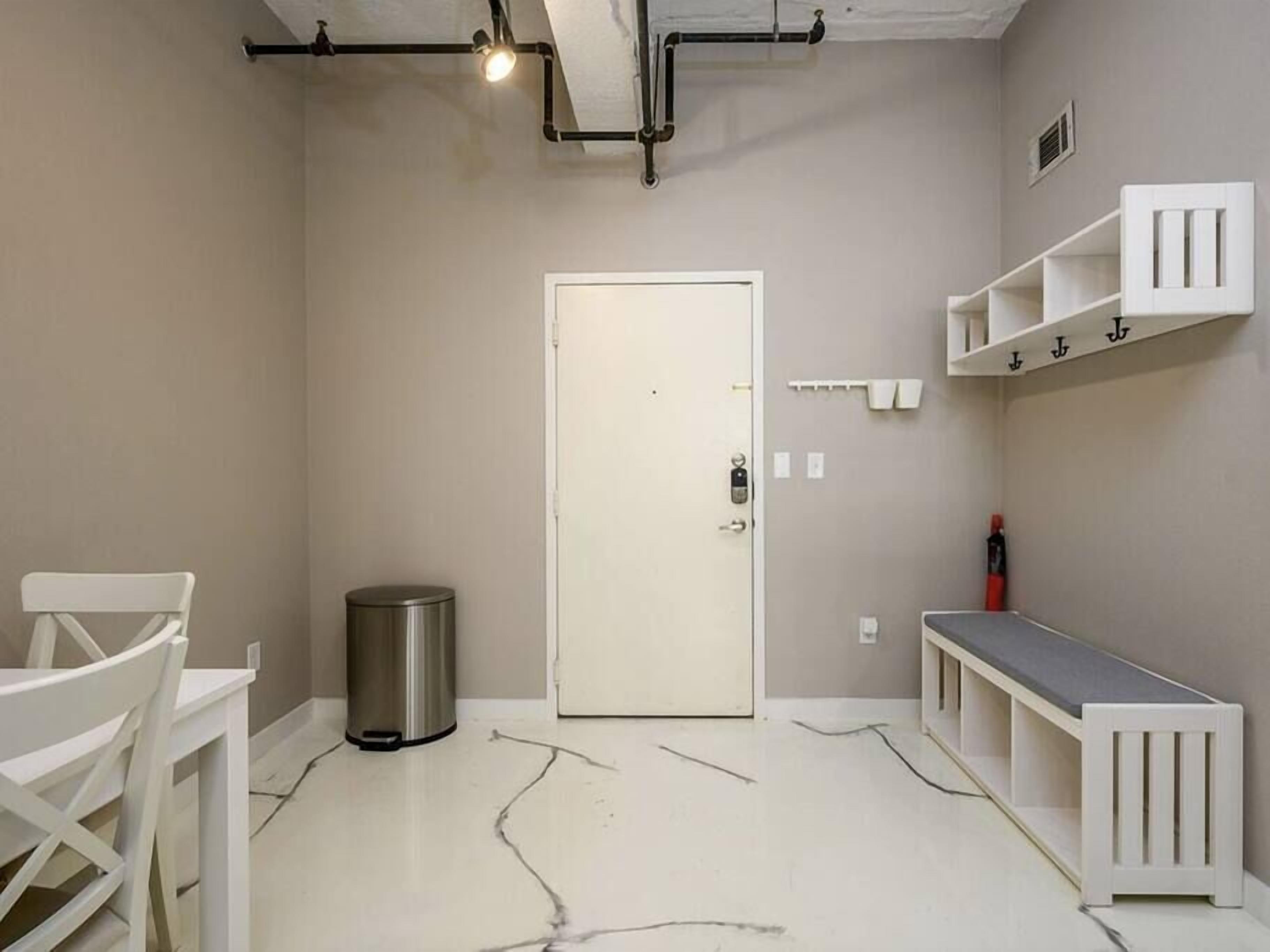 Une pièce avec une porte blanche, une poubelle, une chaise blanche, un banc et une étagère blanche.