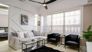 Un salon moderne avec un canapé d’angle, deux fauteuils, une table basse en verre et un tapis noir. De grandes fenêtres sont équipées de stores, et le salon est agrémenté d’un ventilateur de plafond et d’œuvres d’art accrochées aux murs.