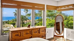 A bedroom with a large wooden dresser, a full-length mirror, and a view of the ocean and palm trees.