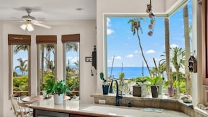 A kitchen with a large window overlooking a tropical landscape, a ceiling fan, and a dining area with chairs.