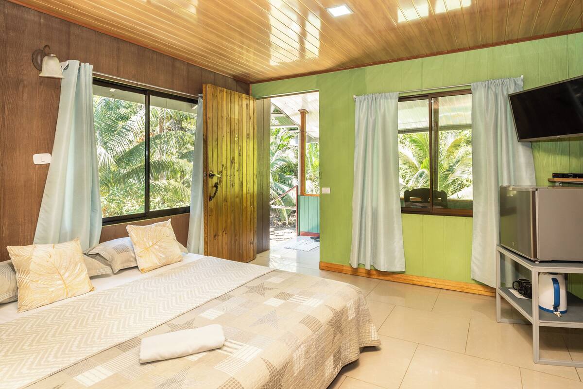 A bedroom with a bed, curtains, a television, and a view of greenery outside.