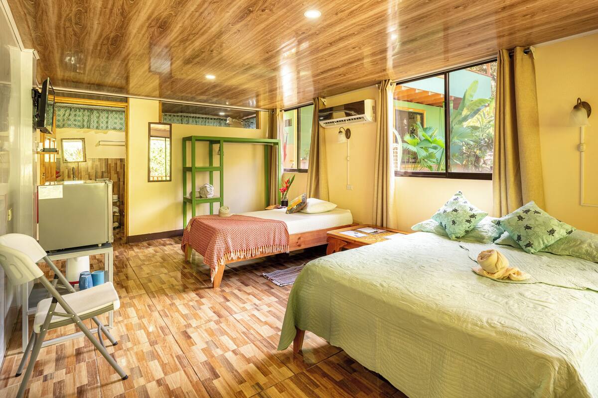 A room with a bed, a desk, and a chair. There is a window with curtains, a ceiling fan, and a wall-mounted air conditioner.