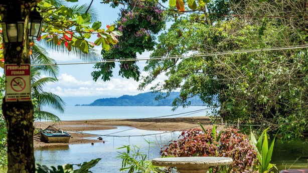 Vista à beira-mar com mesa, cadeiras, uma placa e um barco perto da costa.