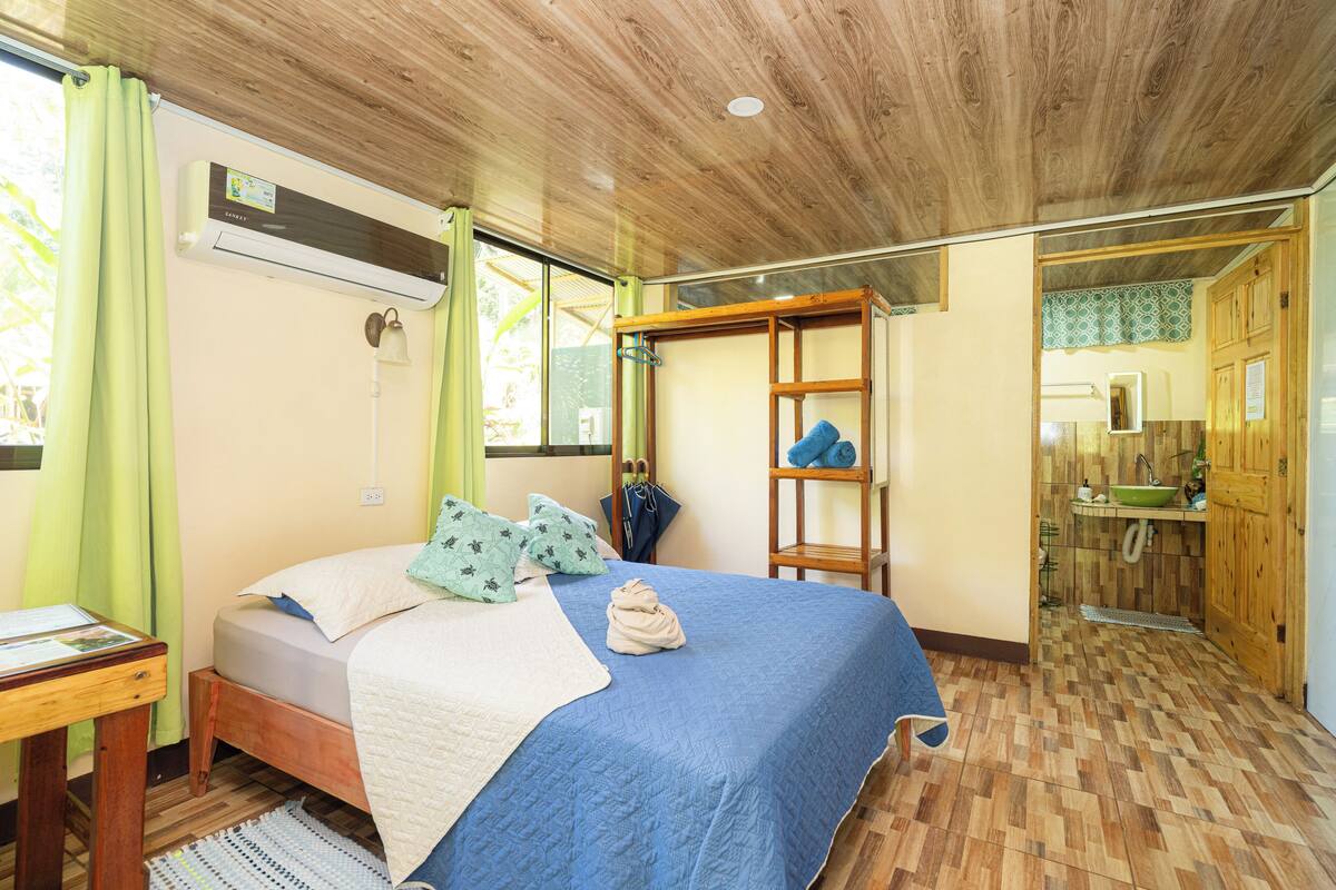 A bedroom with a bed, a wooden ceiling, a window with curtains, a bedside table, and a bathroom visible through an open door.