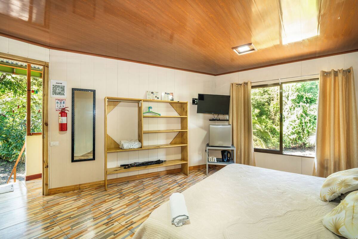 A hotel room with a wooden floor, a bed with a rolled towel, a TV, a shelf with items, and a window with curtains.