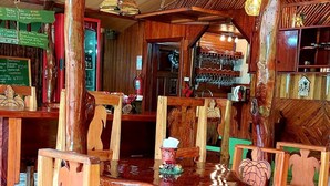 Um interior rústico de madeira com uma mesa redonda central rodeada por cadeiras, um espaço de bar com copos pendurados e um quadro de menu exibindo pratos.