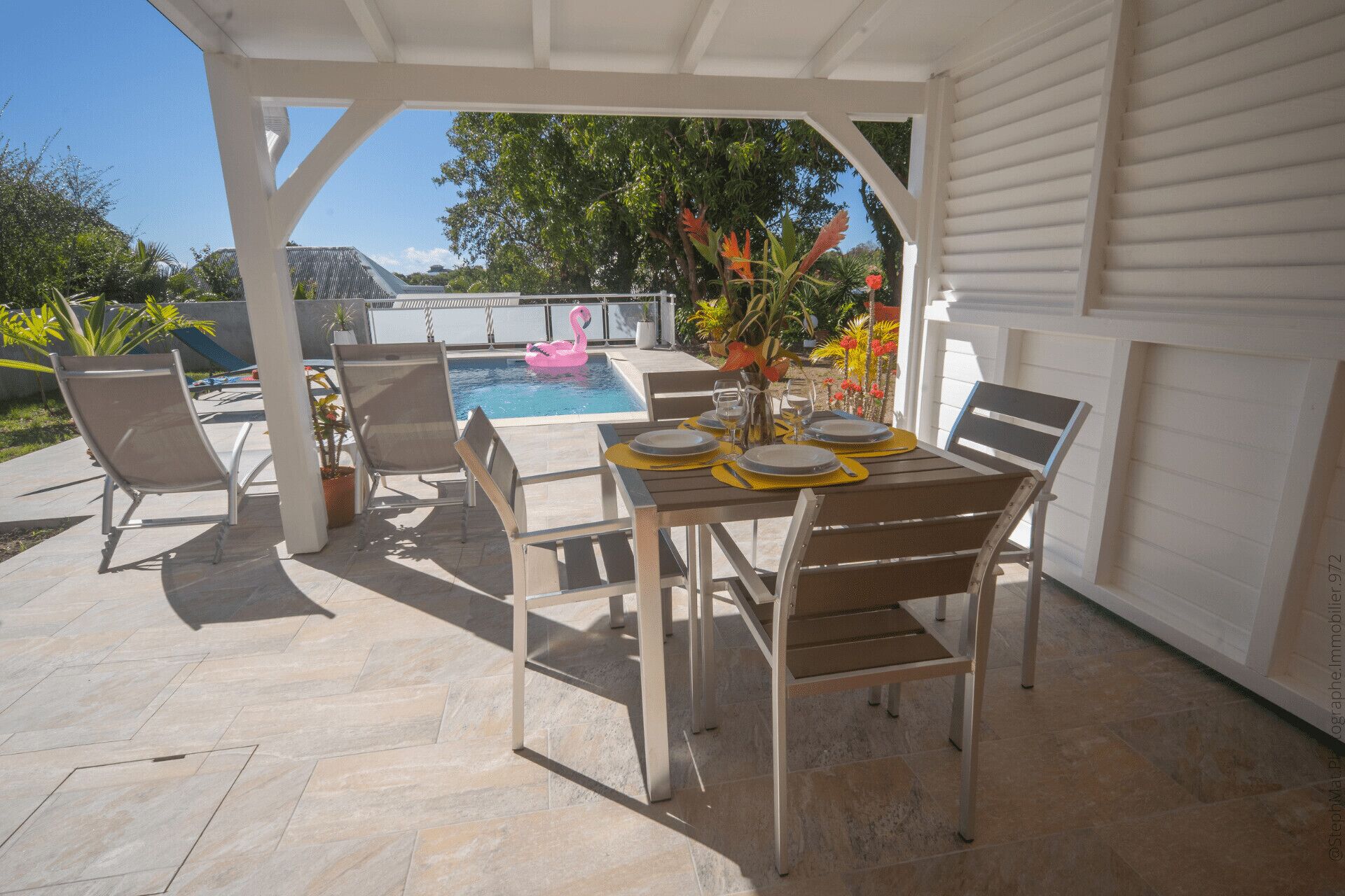 An outdoor patio with a table set for dining, surrounded by chairs, and a pool with a pink flamingo float.