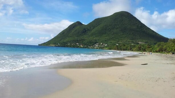 Una playa con aguas azul turquesa, una orilla arenosa y una montaña al fondo.