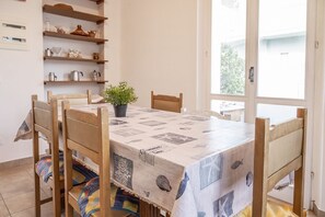 A dining area with a table covered in a patterned tablecloth, wooden chairs, a potted plant, and shelves with kitchen items.