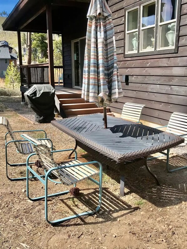 A covered patio with a table and chairs, a striped umbrella, and a wooden house in the background.