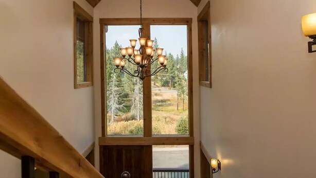 A staircase with a wooden handrail leading to a landing with a chandelier and a large window offering a view of trees and a road. - Castle Cabin - A Spacious Tahoe Retreat. (South Lake Tahoe)