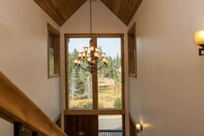 A staircase with a wooden handrail leading to a landing with a chandelier and a large window offering a view of trees and a road.