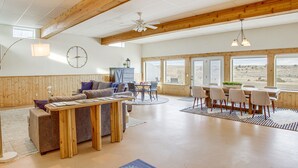A spacious living area with a wooden ceiling, a sofa, a coffee table, and a dining table with chairs. There is a clock on the wall and a view of the outdoors through large windows. - Secluded Montana Getaway w/ Fire Pit & Views! (Ryegate)