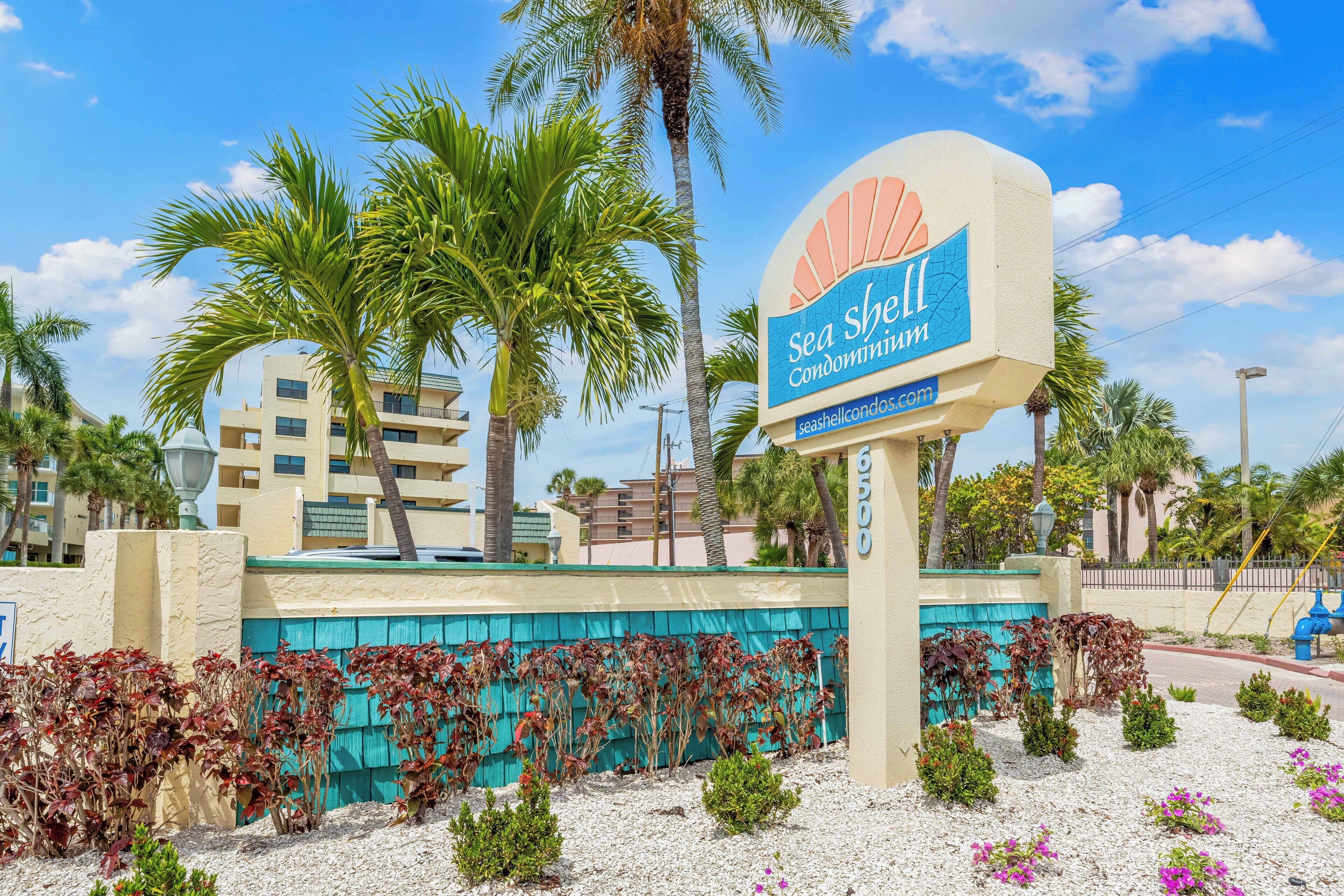 A sign for Sea Shell Condominium with a website address, surrounded by palm trees and a blue fence.