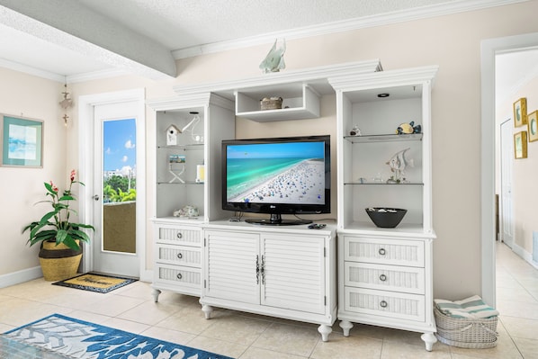 A living room with a television displaying a beach scene, white cabinetry with multiple drawers, a wicker basket, and a potted plant.