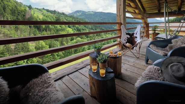 A wooden deck with a view of a forested valley and mountains. There are two chairs and a table with drinks and a plant.
