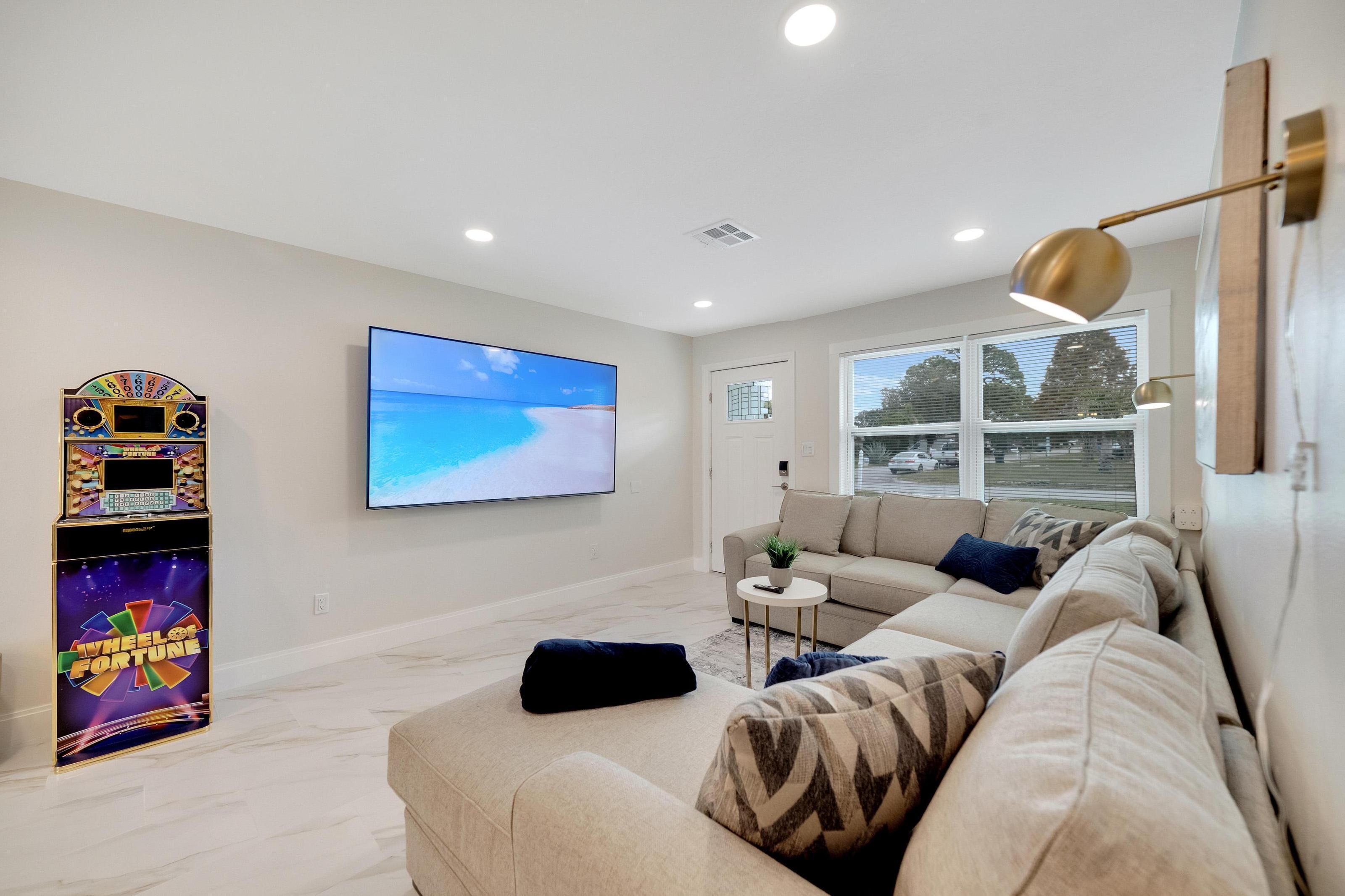 Una sala de estar con un sofá beige, un televisor montado en la pared, una mesita auxiliar y un ventanal con vistas al exterior.
