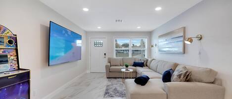 A modern living room with a large flat-screen TV, a beige sectional sofa, a small round coffee table, and a large abstract painting on the wall.