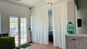 A room with a bed, a green cushion, a white curtain, a teal birdcage, and a window with a view of buildings.