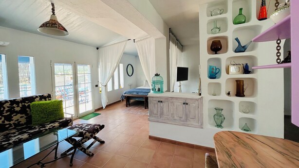 A spacious room with a sofa, a dining table, a cabinet with shelves, and a large window.