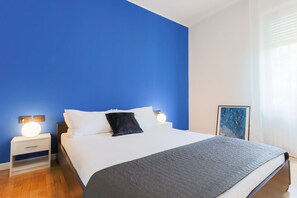 A bedroom with a bed, two bedside tables, a nightstand, a framed picture, and a window with curtains.