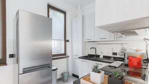 Une cuisine moderne dotée d’un réfrigérateur en acier inoxydable, de meubles blancs, d’un plan de travail noir et d’une fenêtre avec des stores.