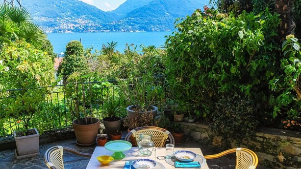 An outdoor dining area with a table set for two, surrounded by potted plants and overlooking a lake with mountains in the background. - Lost in Paradise - My Home In Como (Santa Maria Rezzonico)