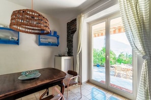 A dining area with a table, chairs, a wicker hanging light fixture, a stone wall, and a view through a glass door to an outdoor patio. - Lost in Paradise - My Home In Como (Santa Maria Rezzonico)
