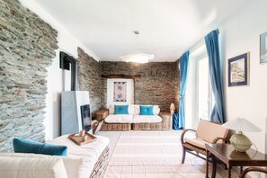 A room with stone walls, a bed with white bedding, a wicker sofa with blue cushions, a wooden table with a lamp, and a framed picture on the wall.