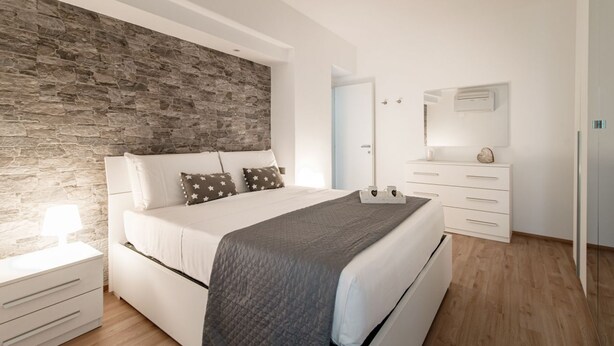 Quarto moderno com parede em pedra, cama grande com manta acolchoada, mesa de cabeceira com abajur, cômoda com gavetas e espelho na parede.