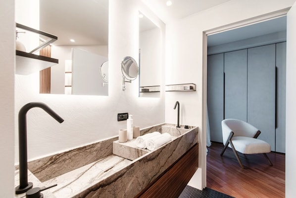 Uma casa de banho moderna com lavatório de mármore, torneira preta e uma cadeira branca na sala adjacente.