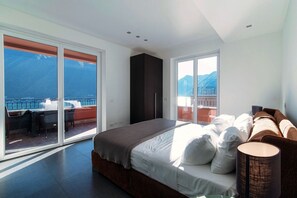 Quarto moderno com cama grande, guarda-roupa e varanda com vista para o mar e as montanhas.
