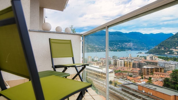 Un balcon avec deux chaises vertes, une vue sur un paysage urbain et des montagnes, et un ciel dégagé.