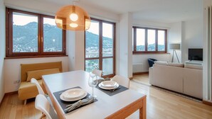 Un salon moderne avec une table à manger pour deux personnes, un canapé et de grandes fenêtres donnant sur un paysage montagneux.