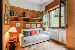 A cozy room with a white sofa, wooden shelves, a table with a lamp, and a window with a view of greenery.