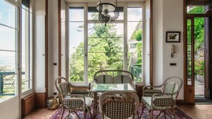 A dining area with wicker chairs, a glass table, and a view of greenery through large windows. - Fairy Tale - I Tigli - My Home In Como (Brunate)
