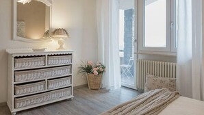 A bedroom with a white dresser, a mirror, a lamp, a wicker basket with flowers, a radiator, and a window with a view. - Wonder View - My Home In Como (Como)