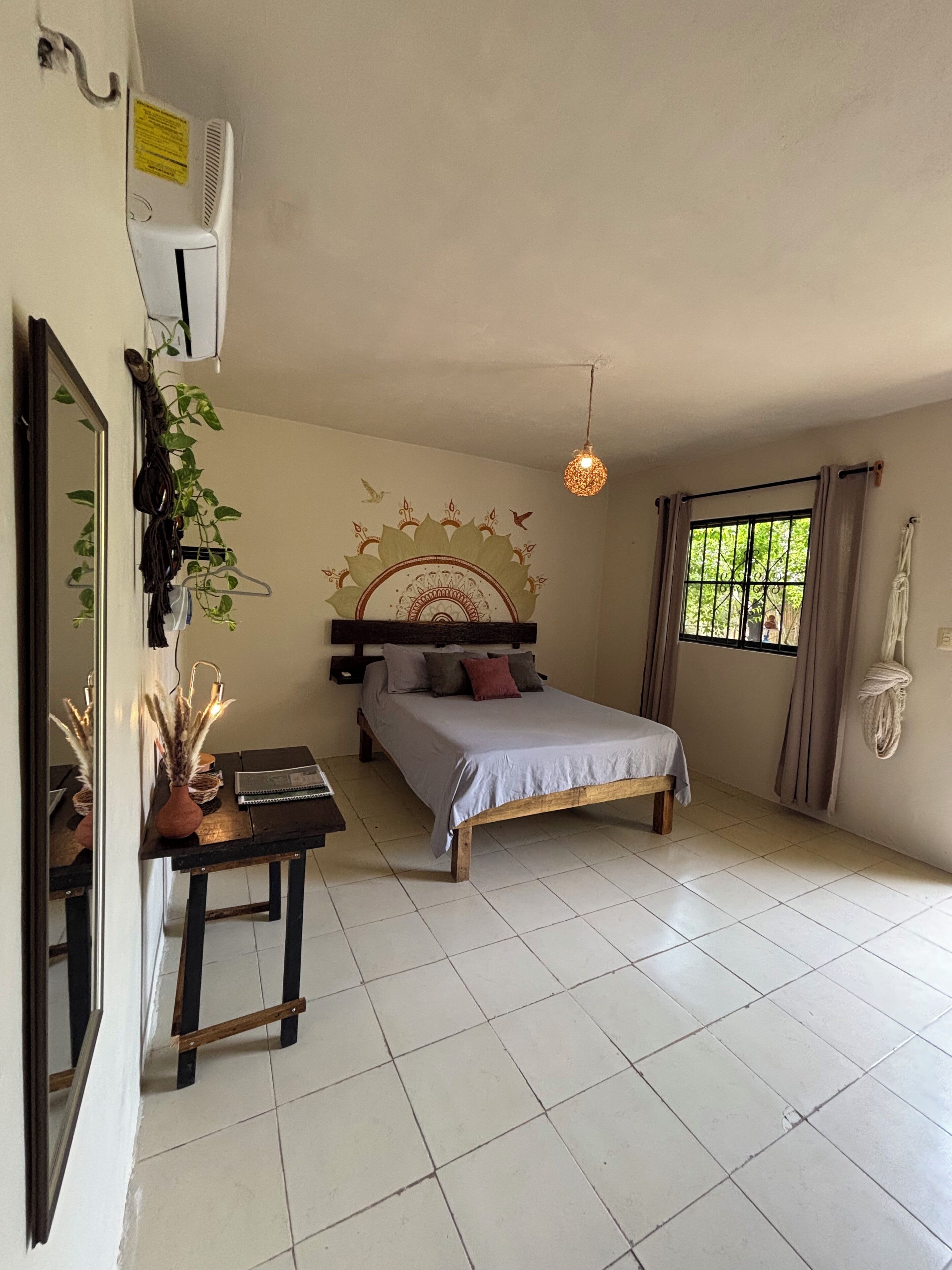 Un dormitorio con cama, mesita de noche, espejo, plantas colgantes y una ventana con cortinas.