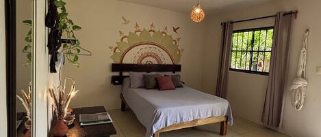 Un dormitorio con cama, mesita de noche, espejo, plantas colgantes y una ventana con cortinas.