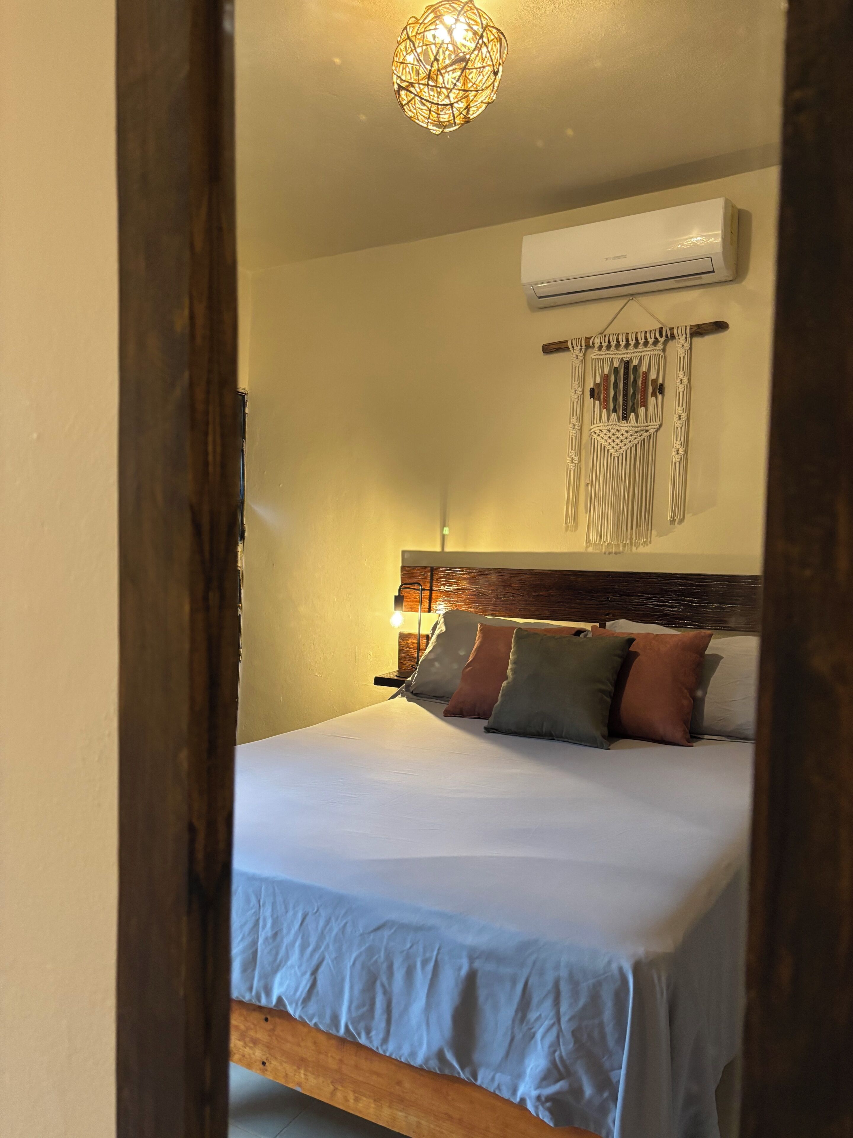 Un dormitorio con cama, aire acondicionado de pared, un colgador de macramé y cabecera de madera.