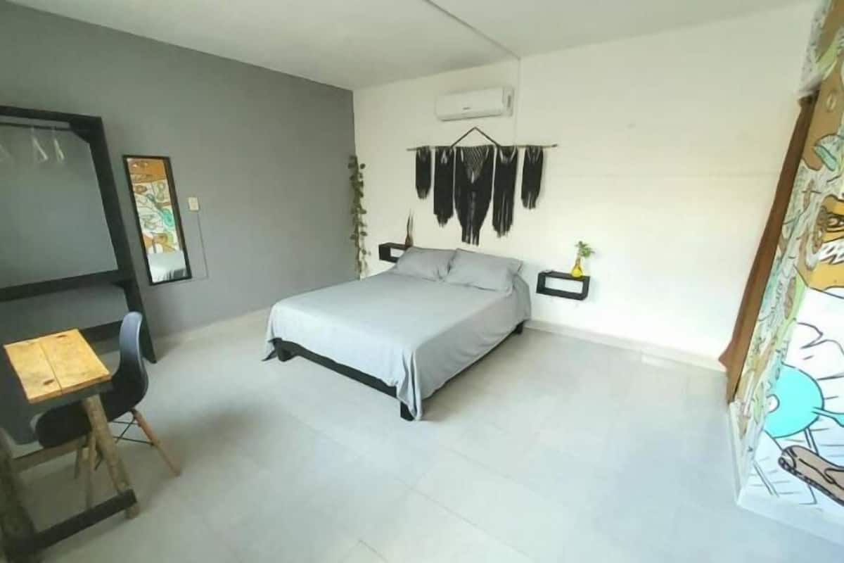 A bedroom with a bed, a desk, and a chair. There is a wall-mounted air conditioner and a framed picture on the wall.